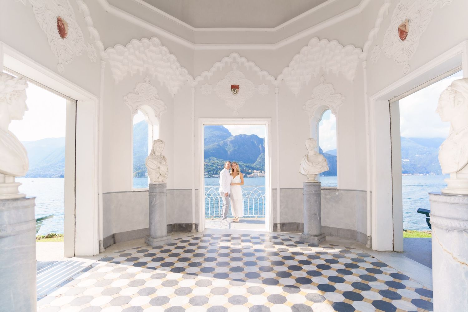 An elegant white colonnade with arched windows and checkered floor tiles facing a mountain landscape.