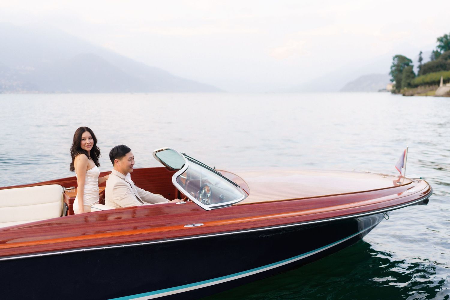 Couple riding in a classic wooden speedboat on a misty lake with mountains in the background.