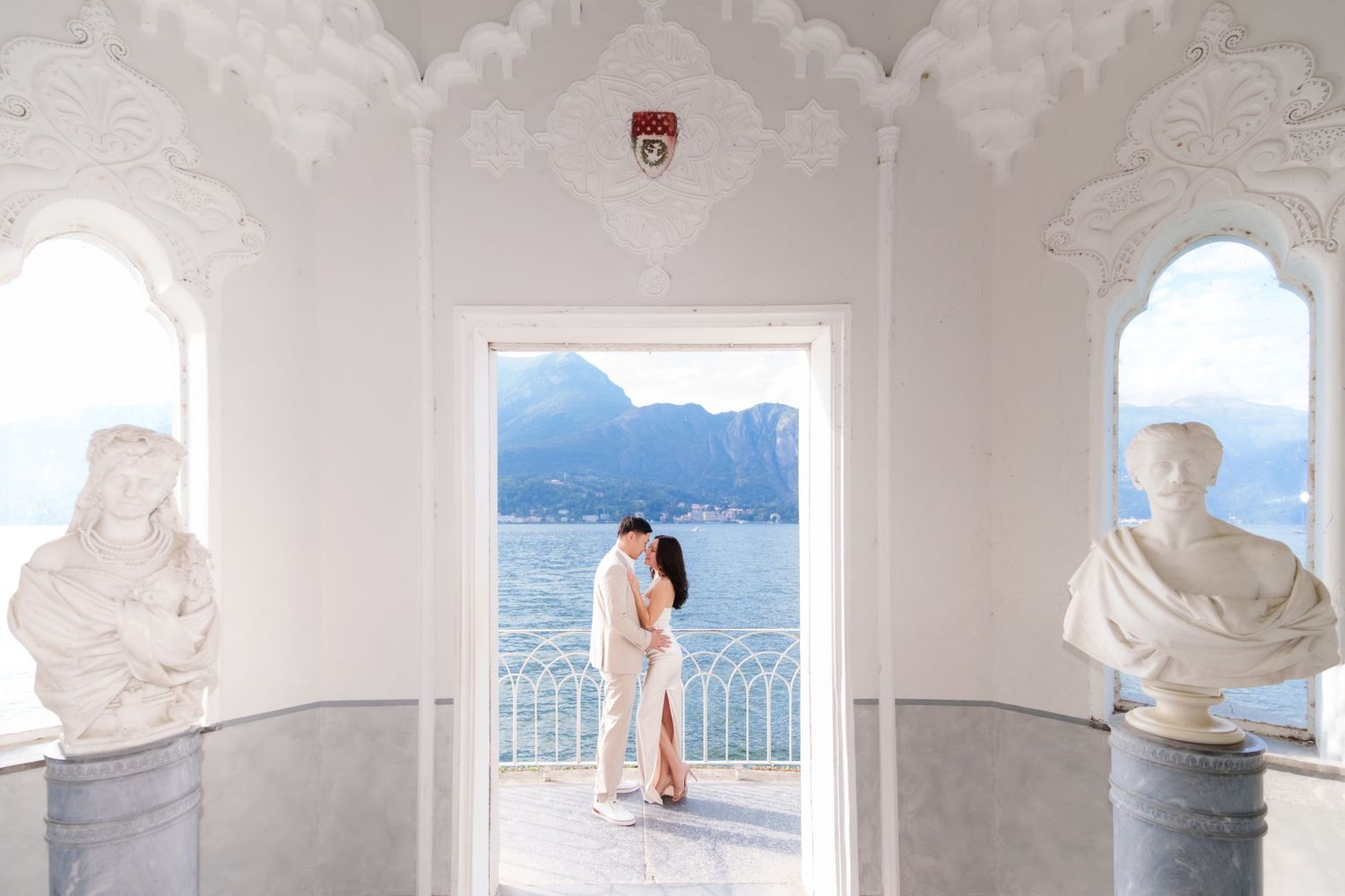 A romantic couple embraces in a white archway overlooking Lake Como in Italy, framed by classical statues and mountain views.