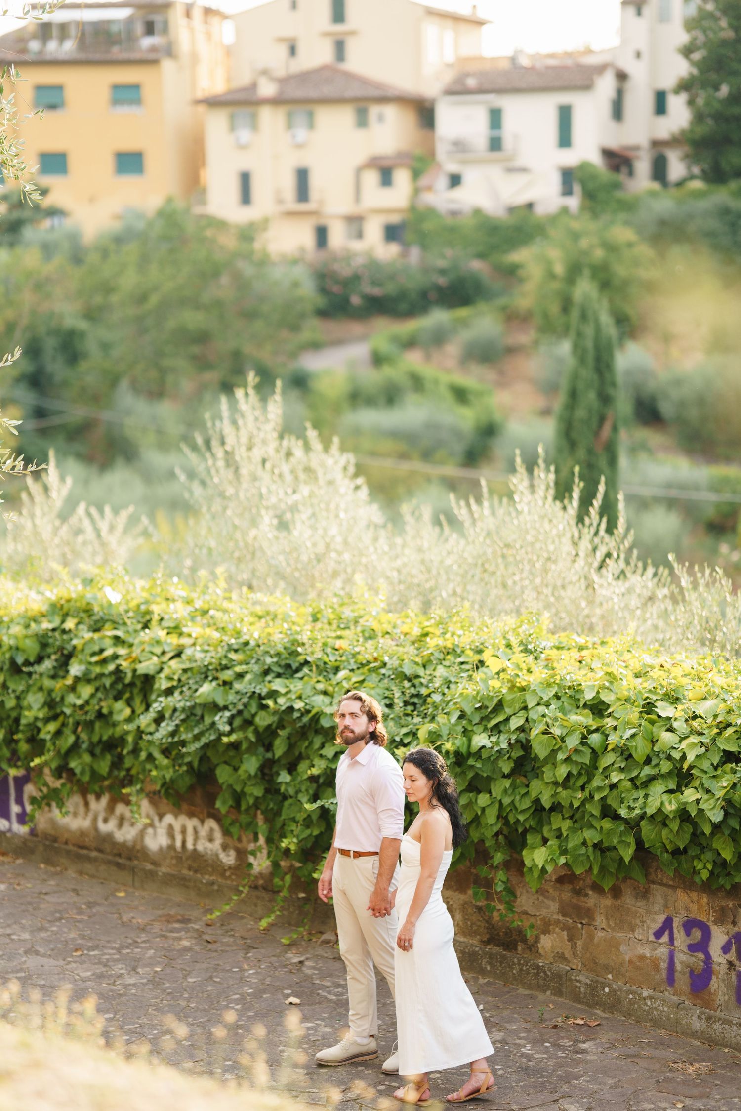 Couple standing on stone path with Italian villa buildings and lush gardens in background during golden hour.