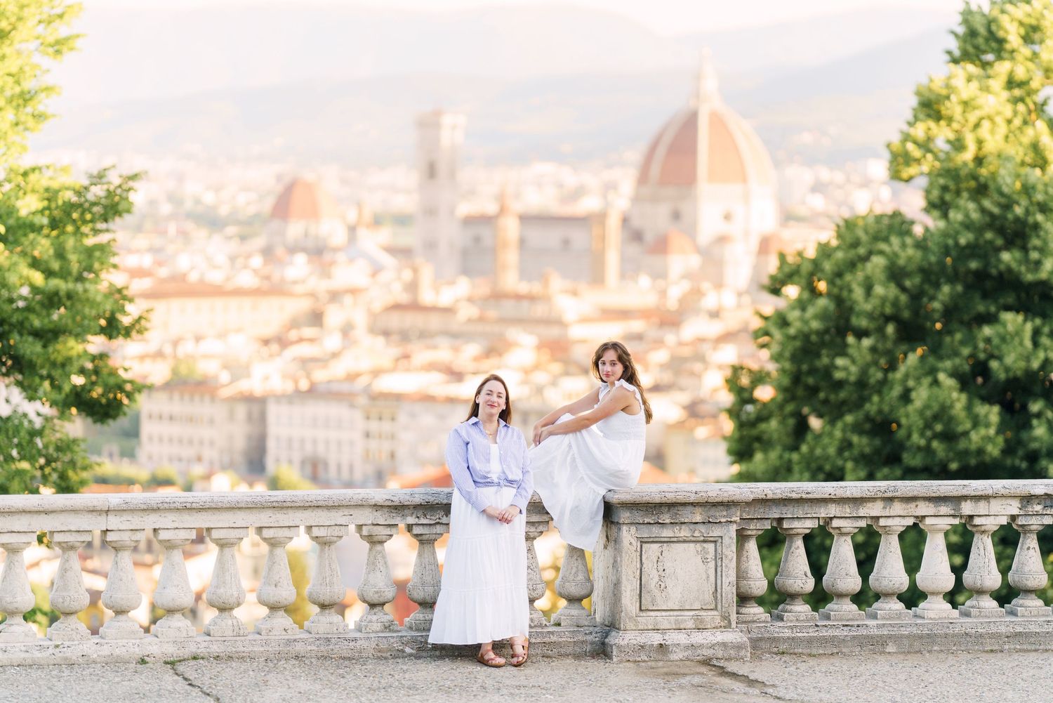 A couple in white outfits poses together on a scenic overlook with a beautiful Italian city vista in the background.