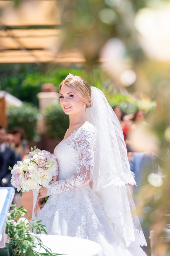 Elegant bride in lace wedding gown carries a pink and white bouquet outdoors.