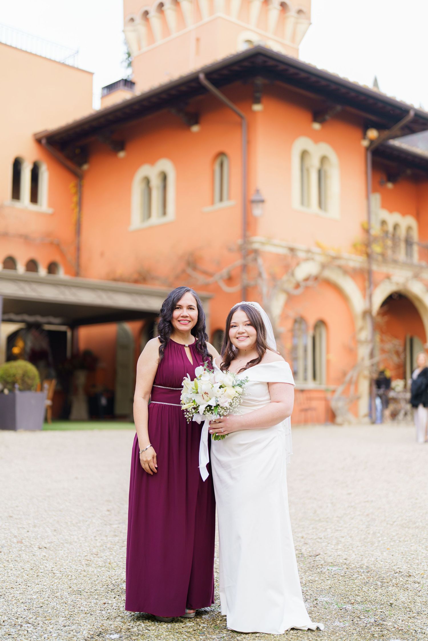 Two people pose together in burgundy and white dresses at a wedding against a Spanish-style building.