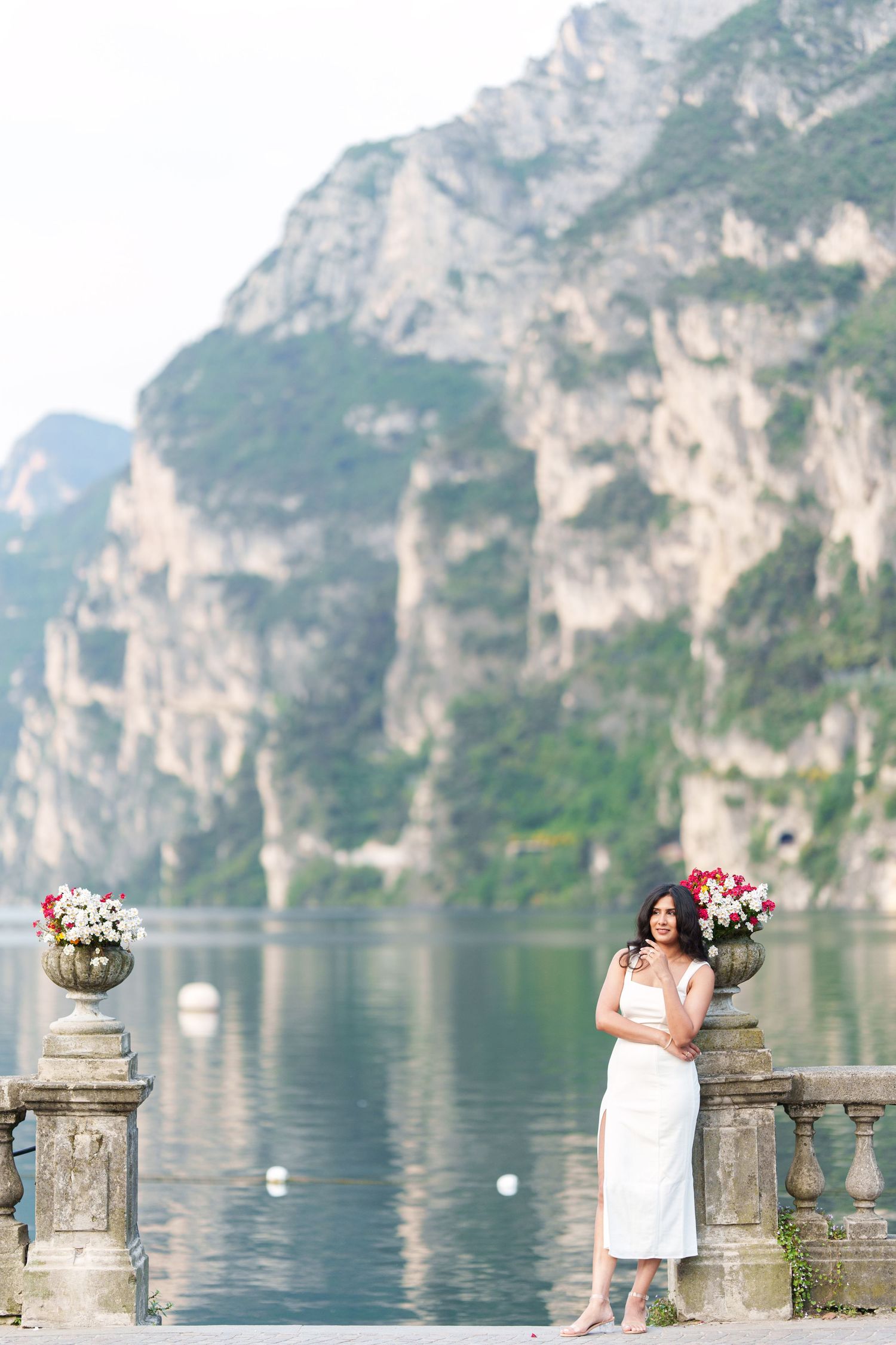 Woman in white dress poses along stone balustrade overlooking scenic Lake Garda with mountain backdrop in Italy.