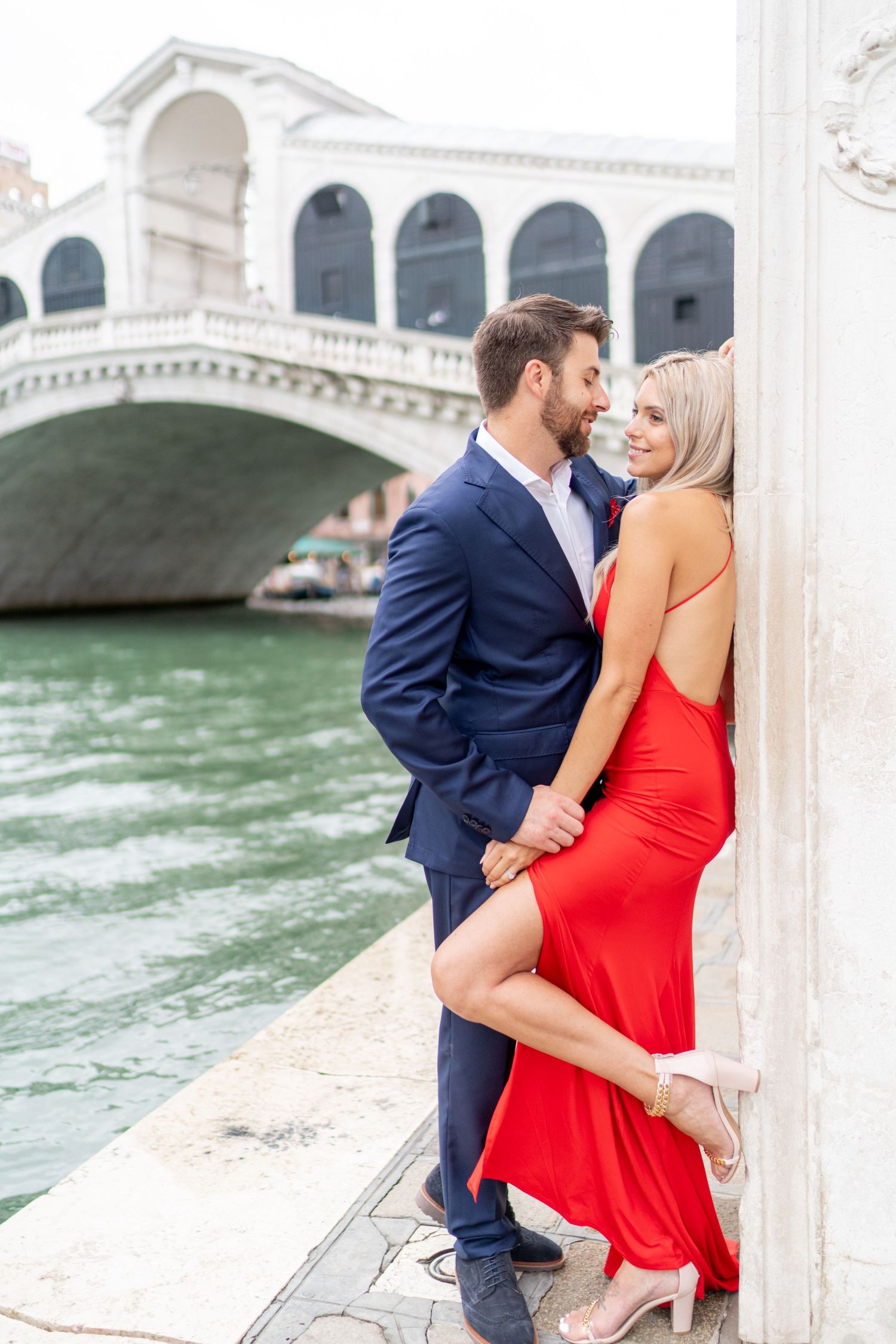 A couple in formal attire shares romantic moments along a canal in Venice with the Rialto Bridge visible in background.