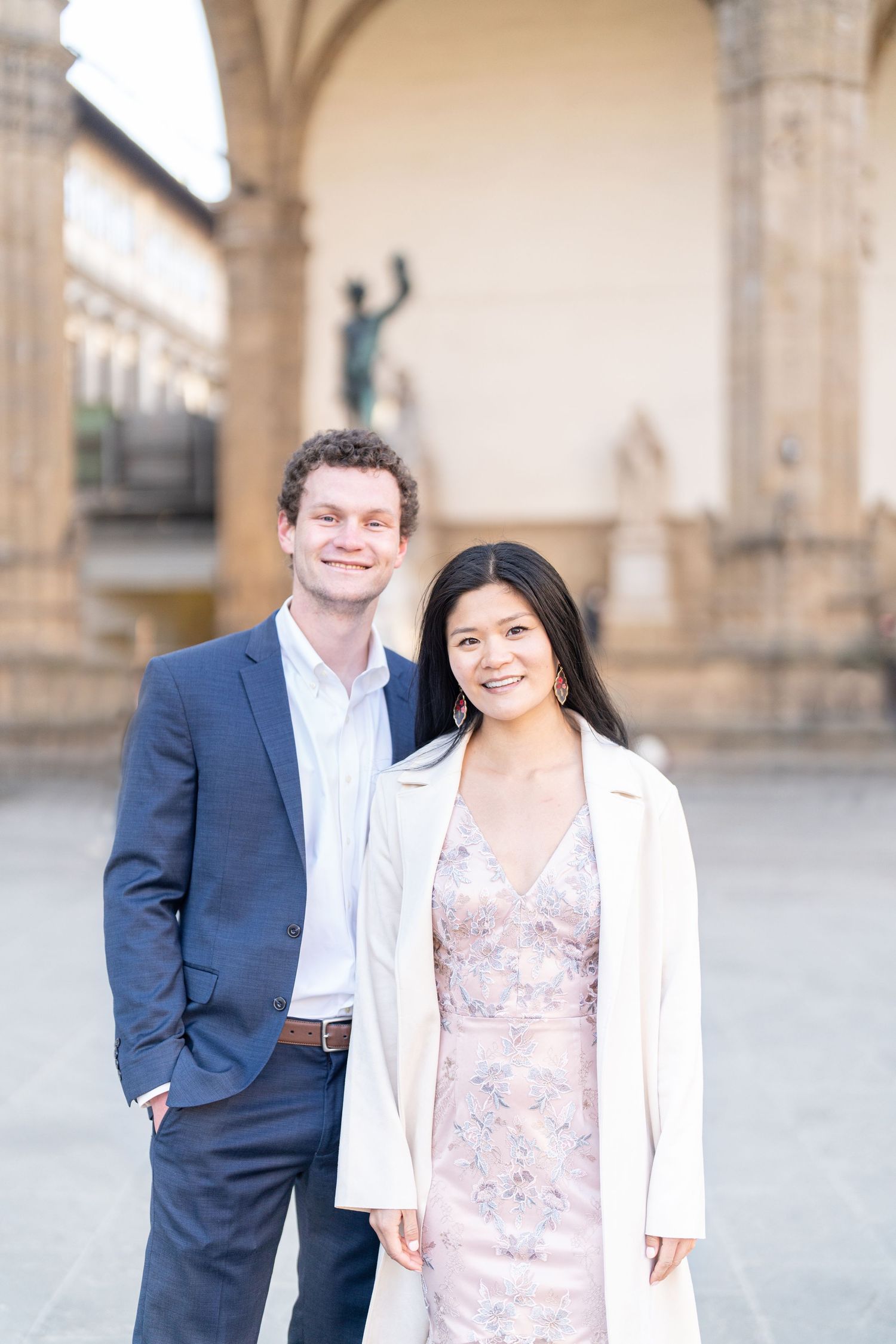A couple poses together under an ornate archway, with a statue visible in the background.