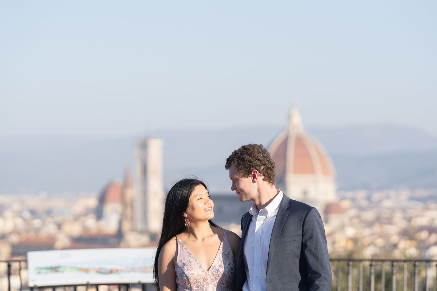 A romantic couple stands on a scenic overlook with a beautiful European city skyline in the background.