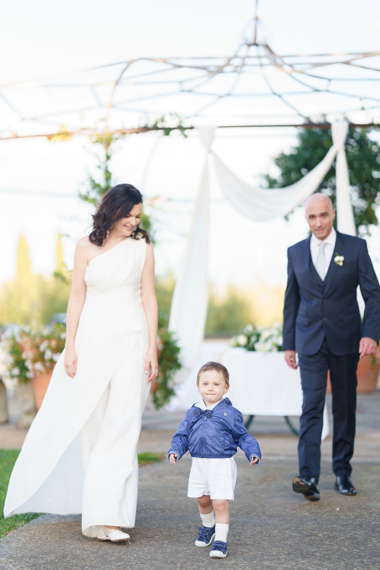 Wedding party walking along a garden path with terracotta planters and white tent canopy overhead.