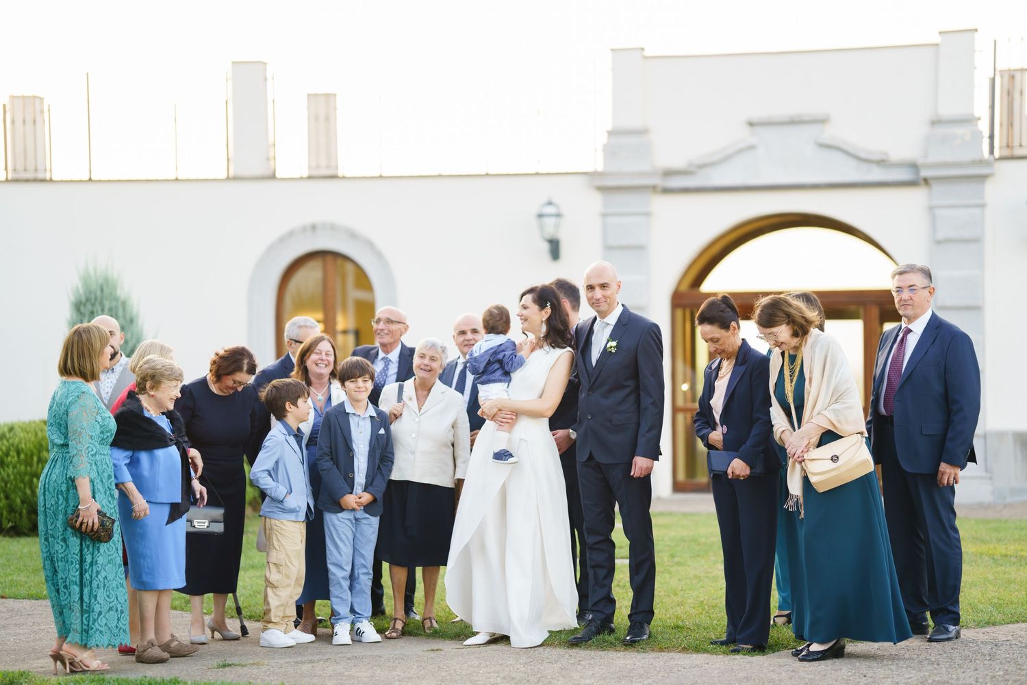 Wedding party and guests gathered outside a white church building for formal group photos.