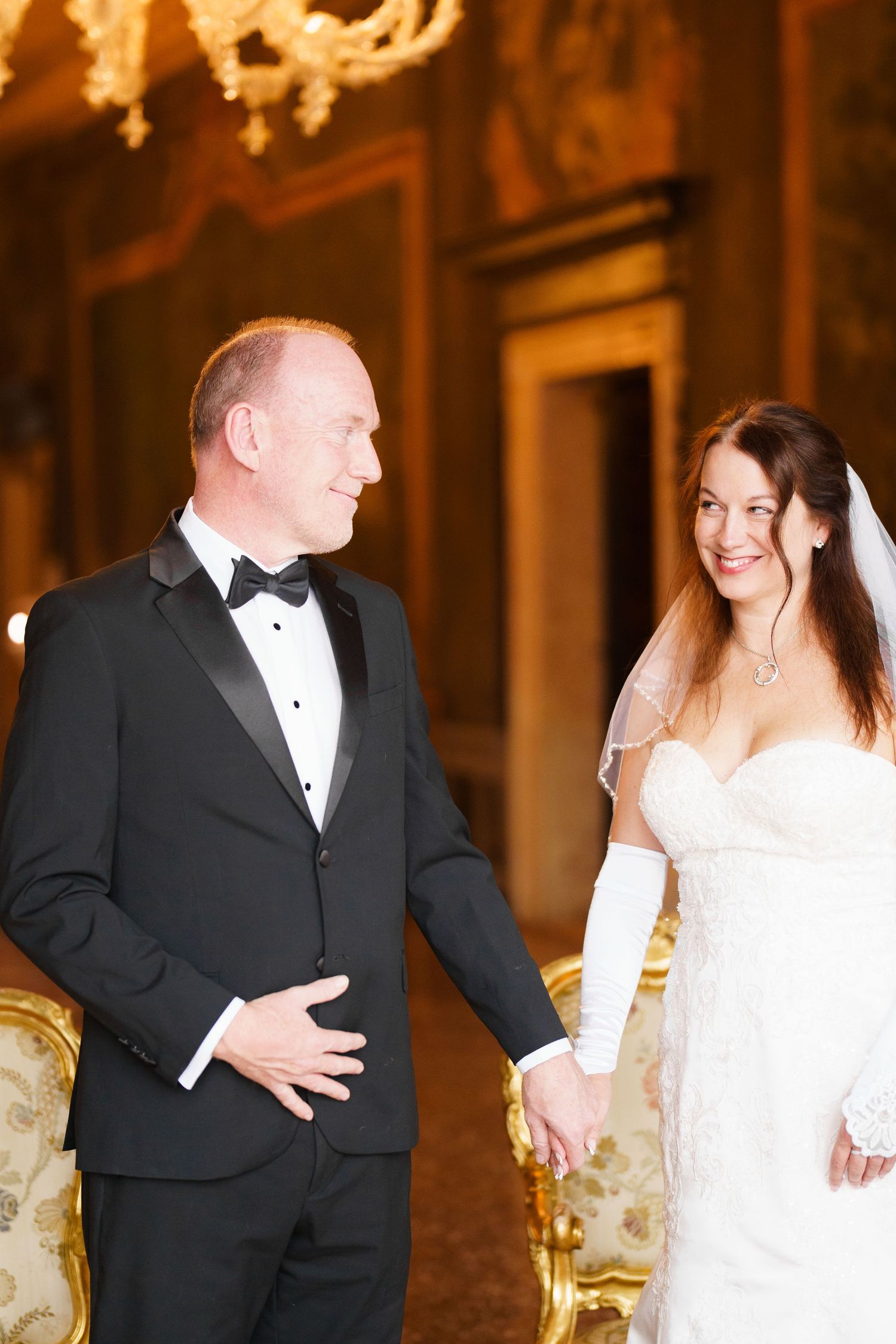 A bride in a white lace dress and groom in a black tuxedo share a moment in an elegant, ornate room.