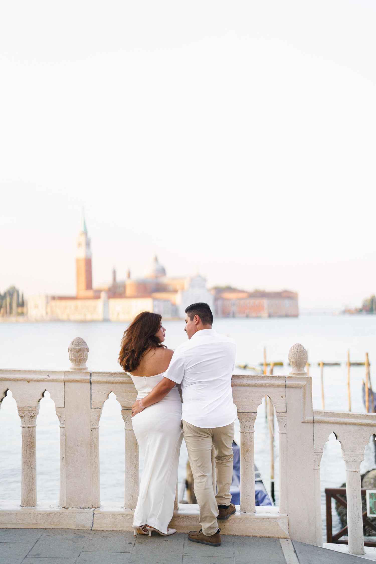 A couple in white attire leans against a stone balcony overlooking a waterfront cityscape during sunset.