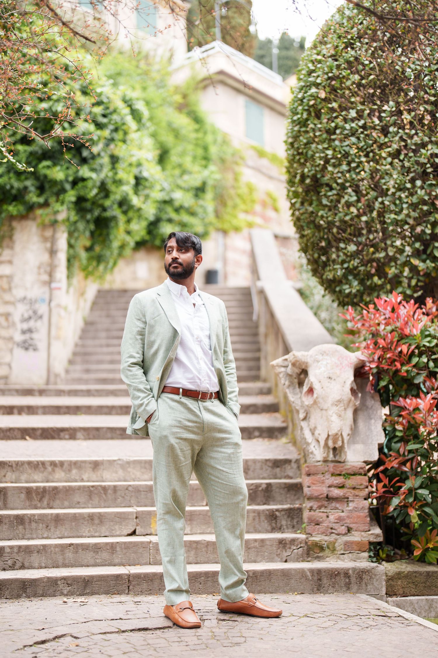 photographer verona A person in a mint green jumpsuit and brown belt poses on stone steps surrounded by lush greenery and flowers.