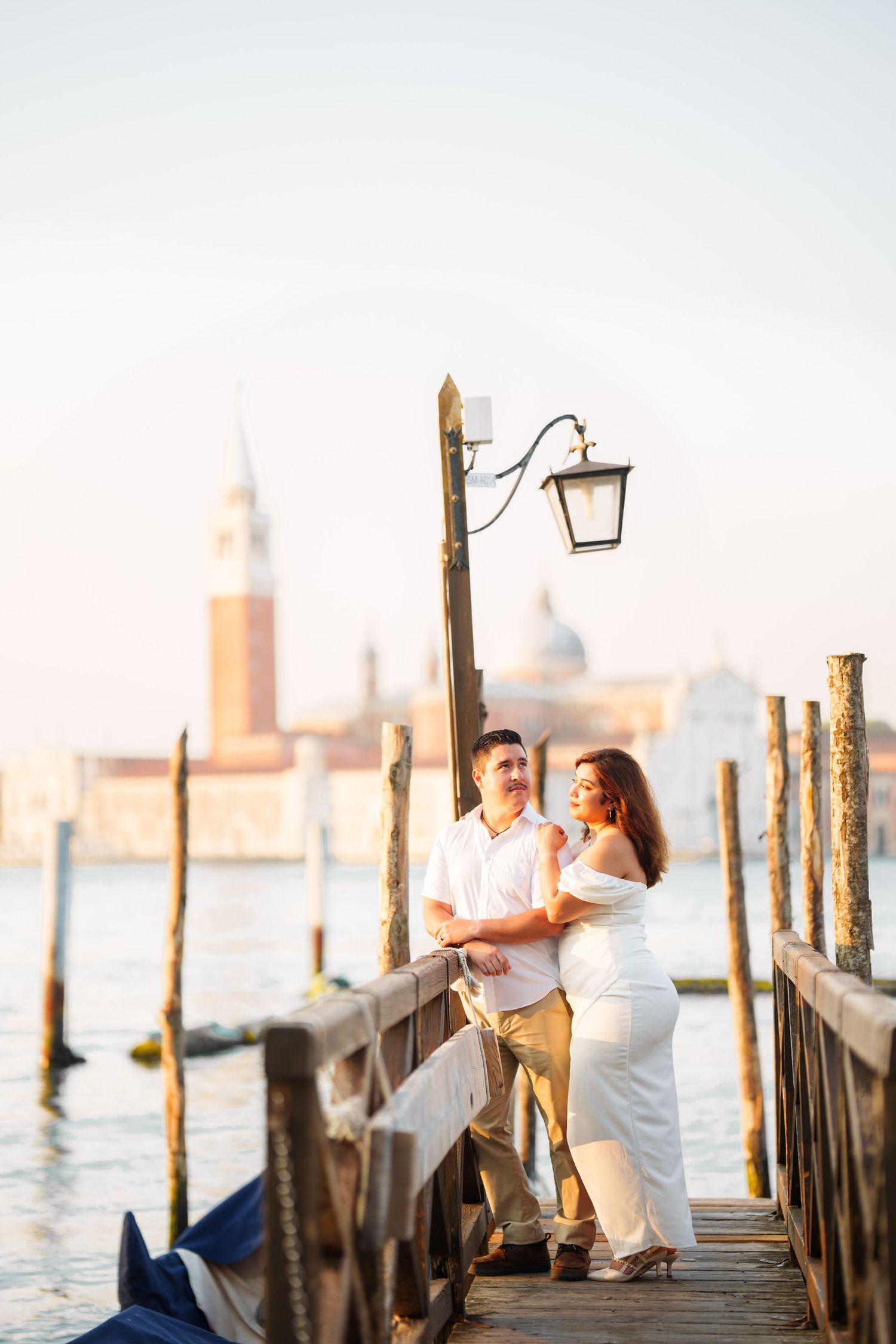 A couple shares a romantic moment on a wooden pier in Venice with the San Giorgio Maggiore church in the background.