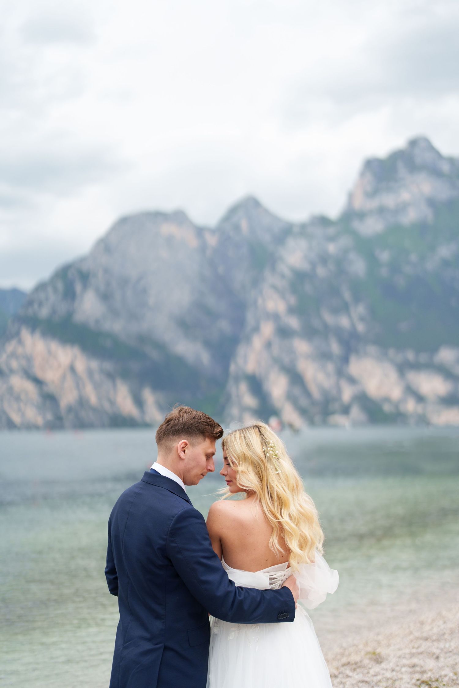 A romantic wedding couple embraces on a beach with dramatic mountain peaks rising from a misty lake in the background.