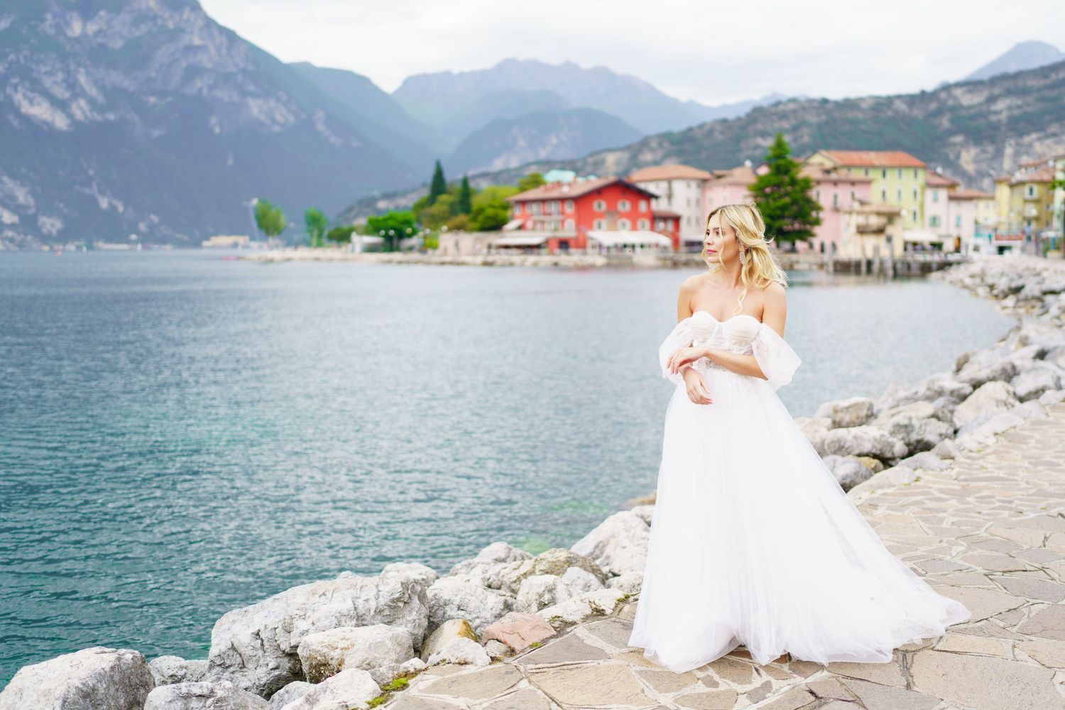 A wedding couple stands along the rocky shores of Lake Garda in Italy with mountains and charming buildings in view.