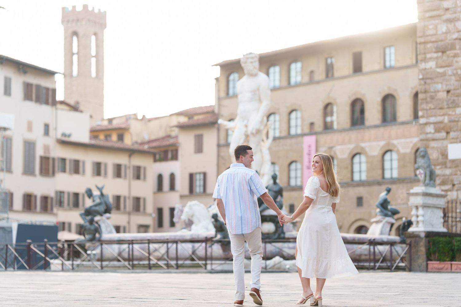 A couple strolls through a picturesque Italian plaza with historic stone buildings and architecture in the background.
