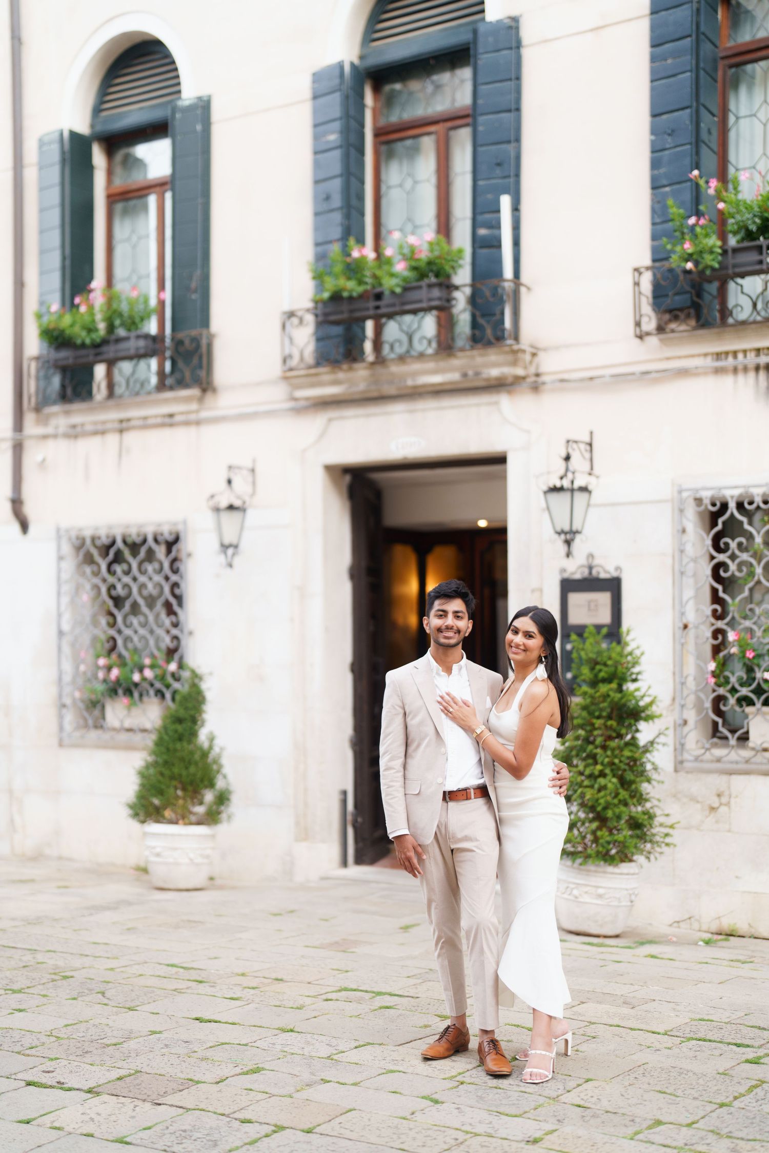 Couple in elegant white outfits poses together in front of historic European building with window boxes and stone facade.