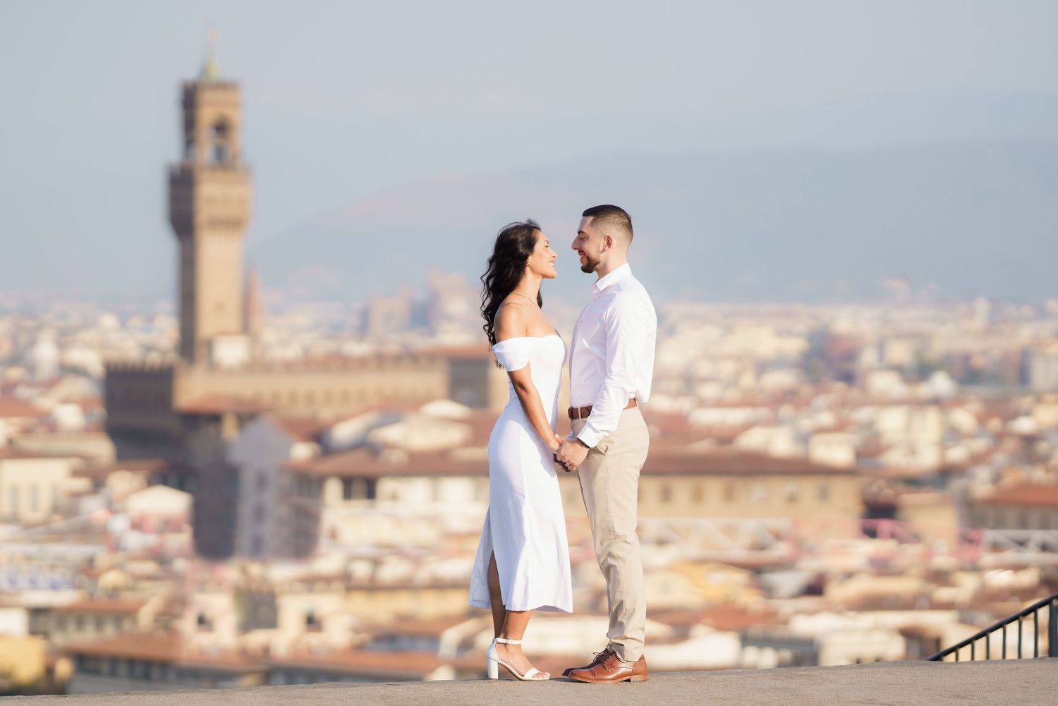 A romantic couple stands on an overlook with Florence's historic cityscape and Palazzo Vecchio tower in the background.