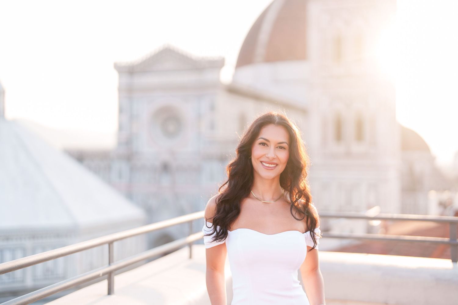 Person in white off-shoulder dress smiles on rooftop terrace with sunlit church domes in background.