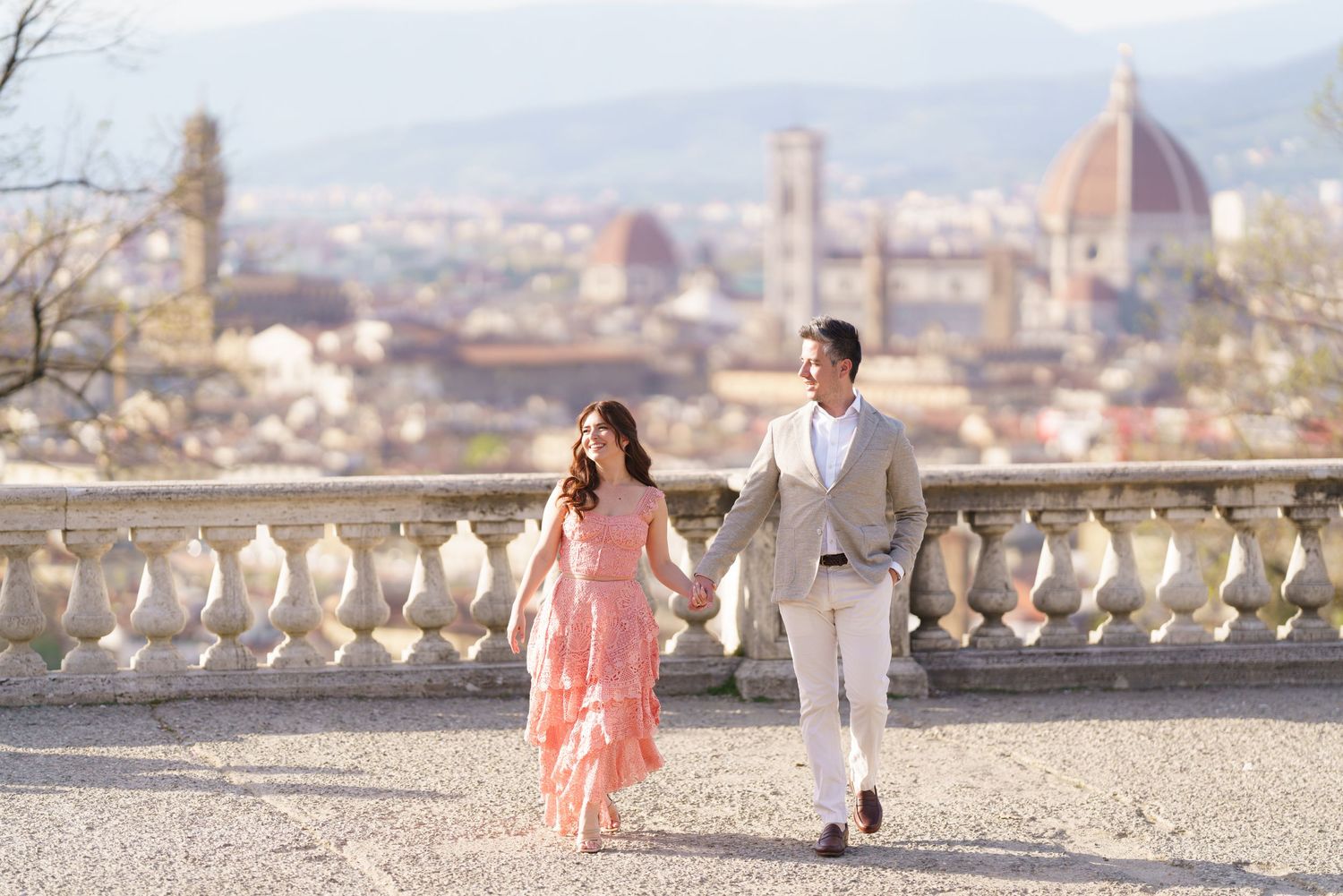 A couple walks along a scenic overlook with Florence's iconic Duomo cathedral and cityscape in the background.