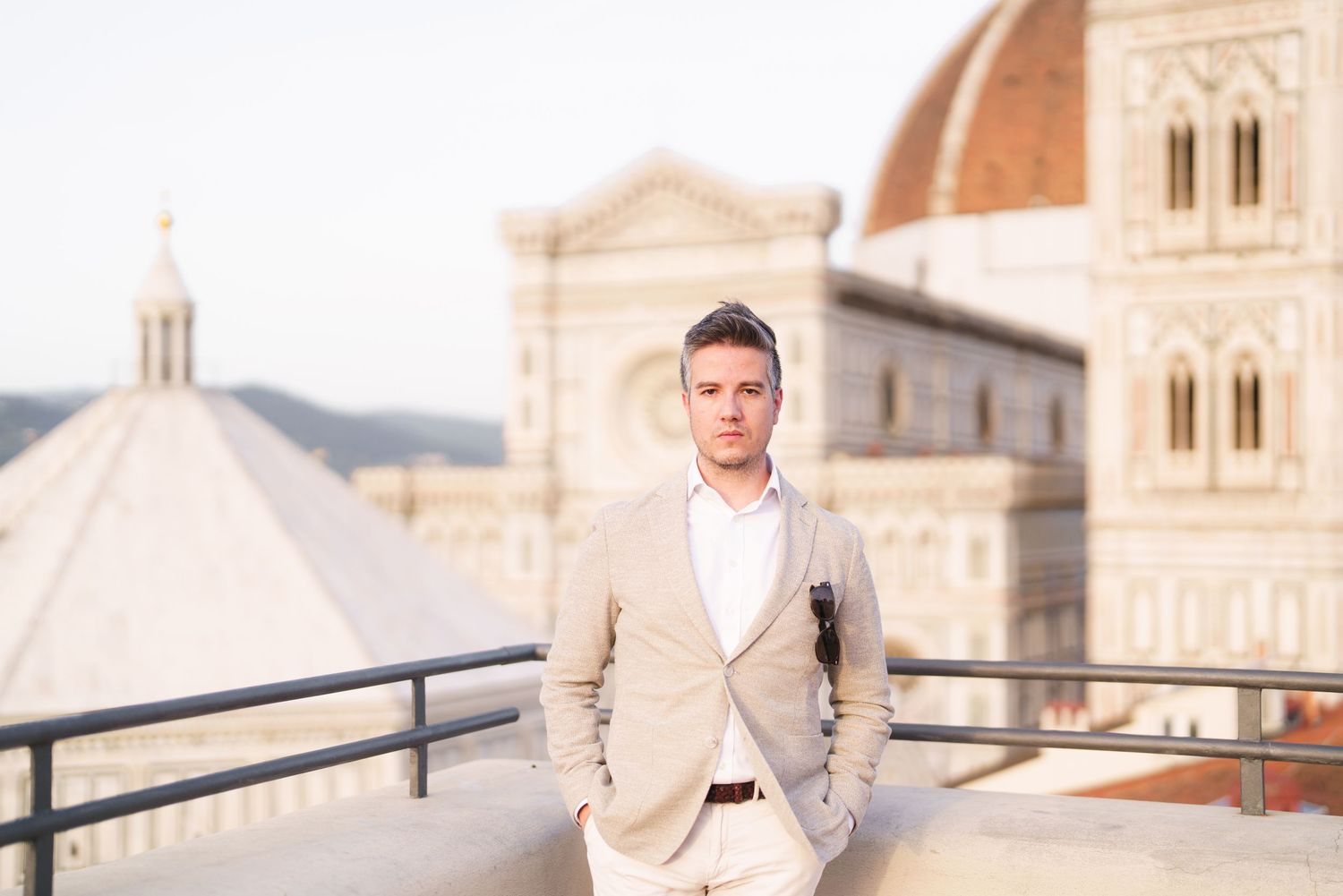 A person in a beige suit poses against a scenic European cityscape backdrop with historic architecture.