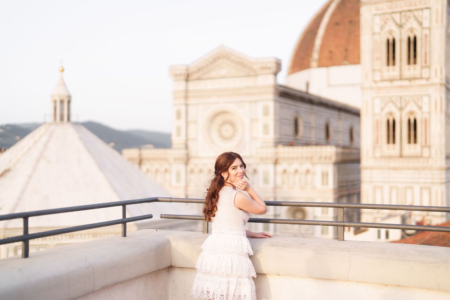 A person in a white dress poses on a rooftop terrace with Florence Cathedral's iconic dome in the background.