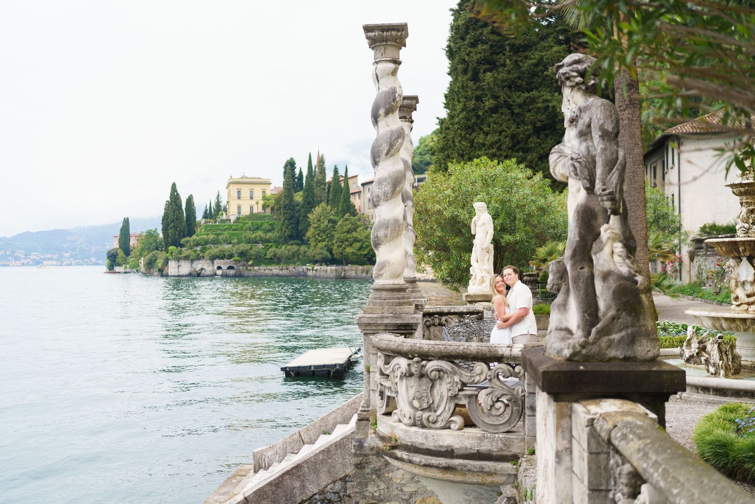 Couple sits among ornate stone statues on a lakeside terrace at Villa Monastero, Lake Como, Italy.