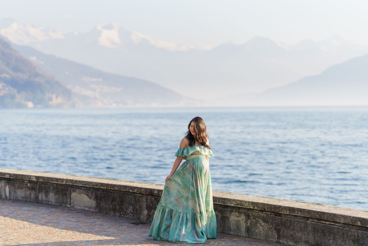 Woman in teal floral gown stands by a stone promenade overlooking a serene lake with misty mountains beyond.