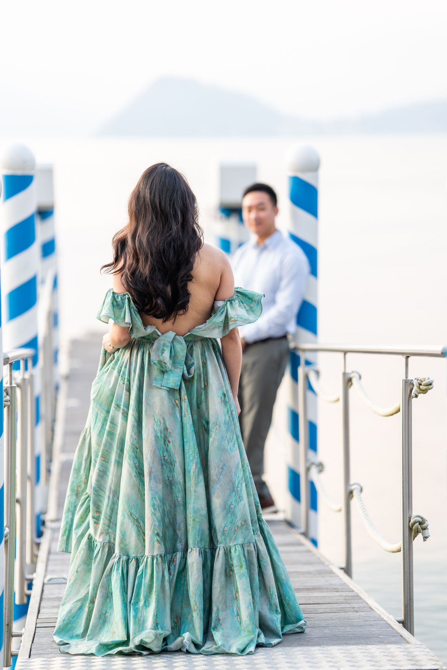 Couple on a pier, woman in elegant green off-shoulder gown with bow, man in blue shirt, ocean and mountains behind.