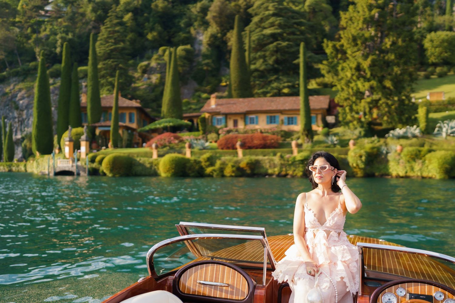 Woman in floral dress on wooden boat at Lake Como with lush greenery and elegant villa in background.