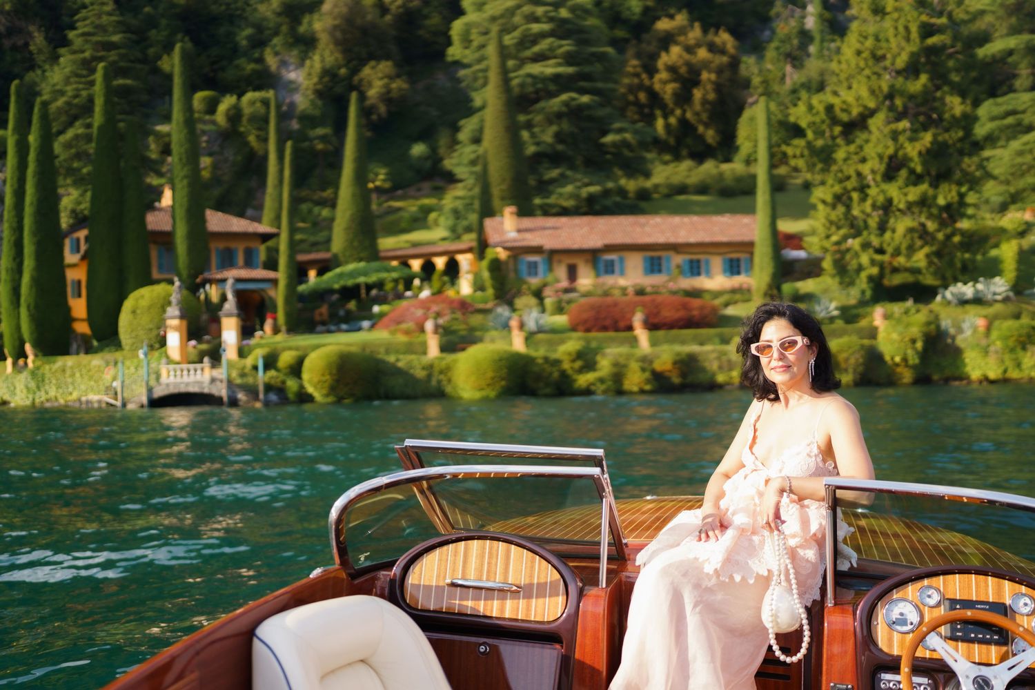 Woman in pink dress sitting on a wooden boat on Lake Como, Italy, with lush green gardens and villa in background.