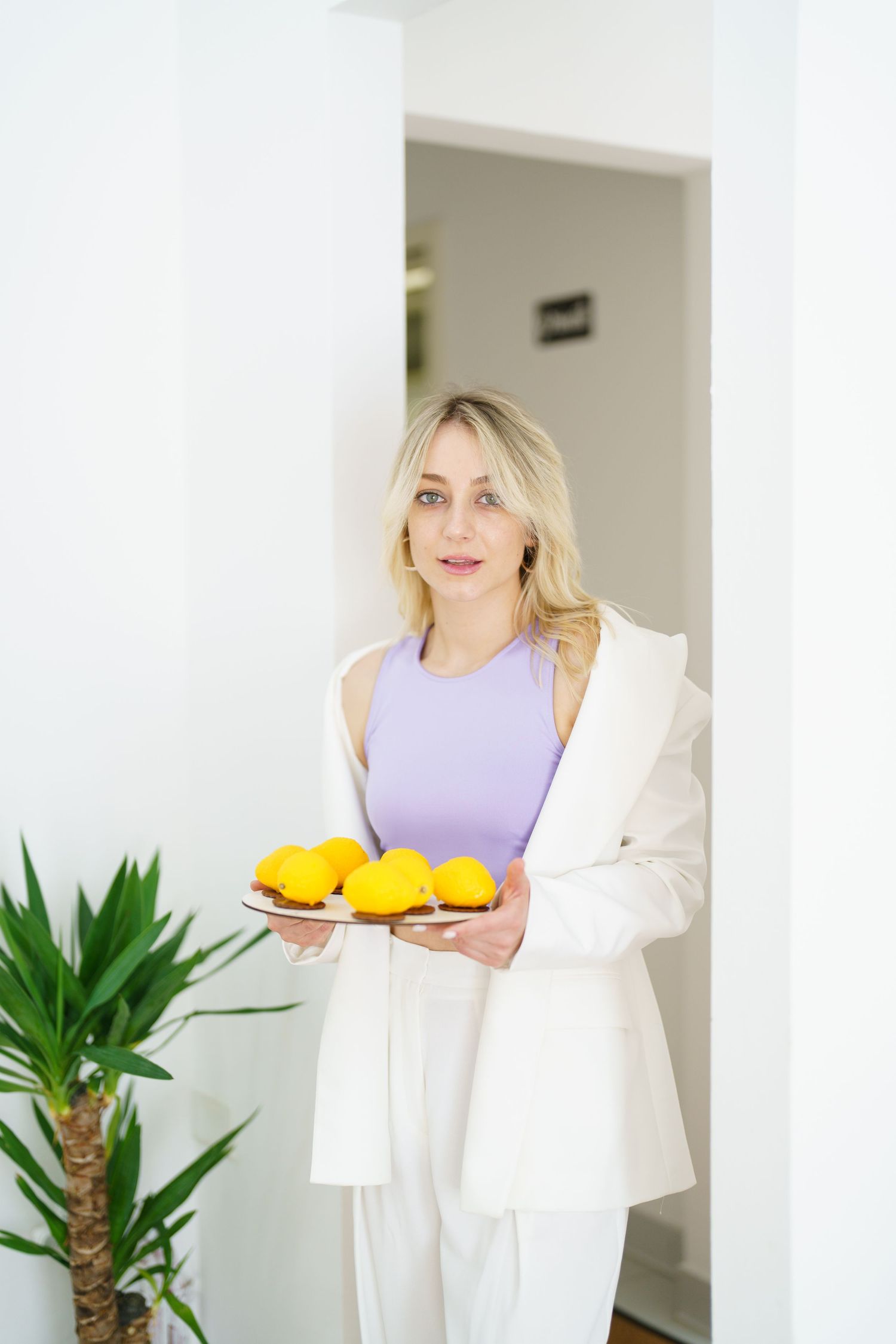 Blonde woman in white blazer and lavender top holding a plate of yellow lemons in a bright white room.