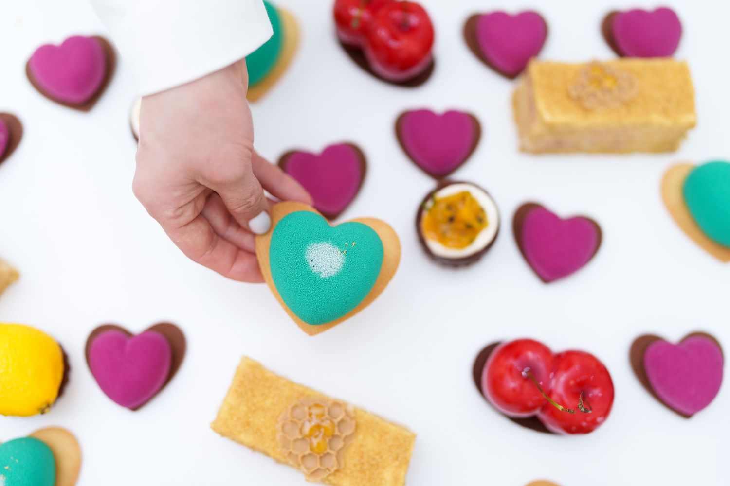 A hand holds a teal heart-shaped cookie among colorful purple, red, and gold heart cookies on a white surface.