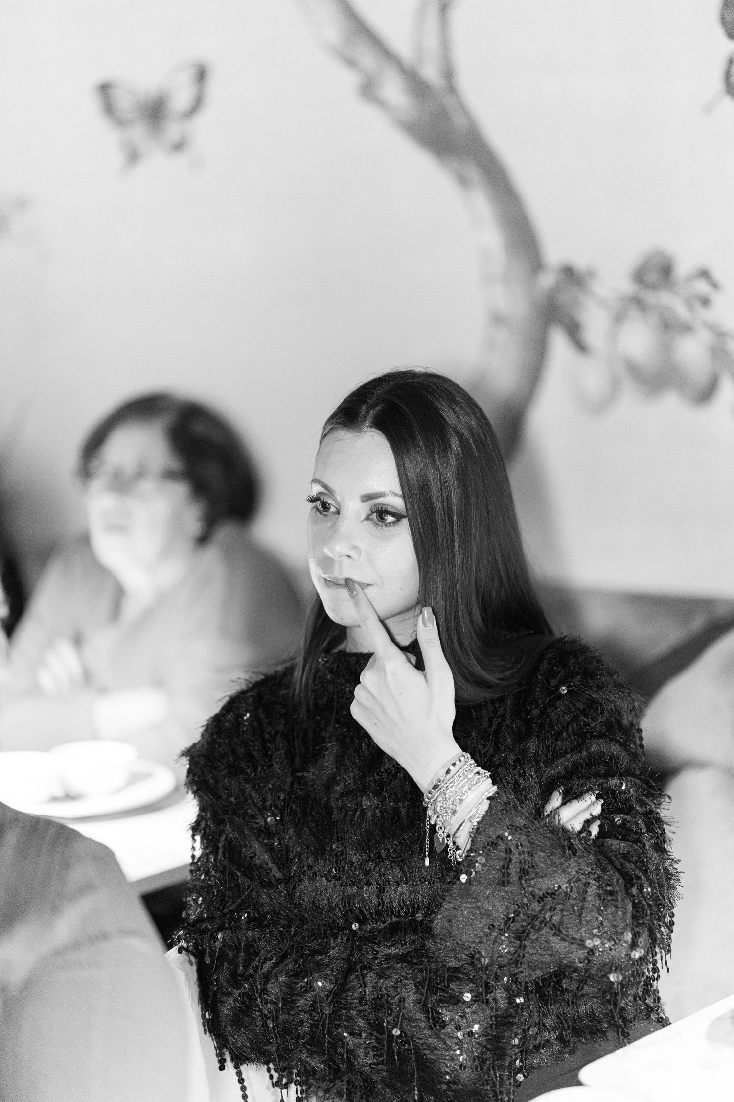 Black and white photo of an elegant woman in a feathered jacket, posing thoughtfully at a restaurant table.