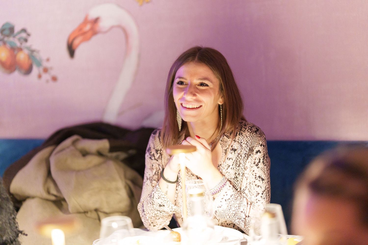 Smiling woman sitting at a candlelit dinner table with a flamingo mural on the pink wall behind her.