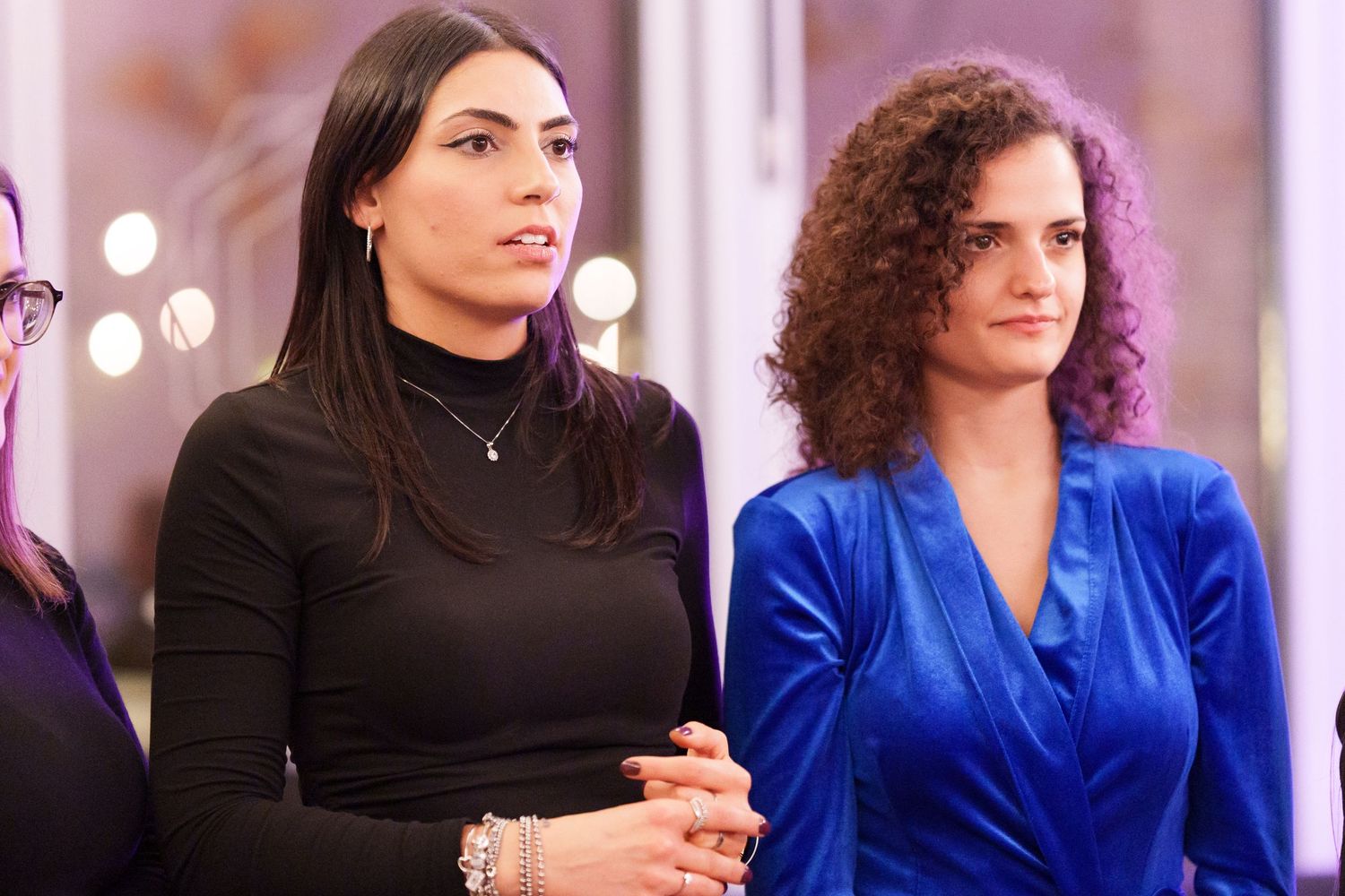 Two women standing together, one in a black turtleneck and one in a blue velvet dress, at an indoor event.