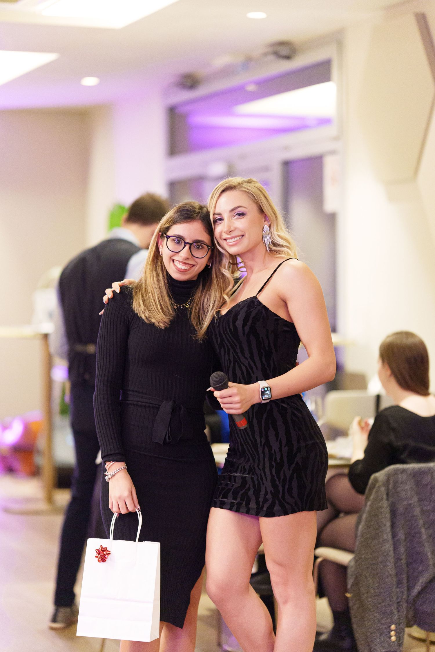 Two women in black outfits pose together at an indoor event, one holding a gift bag and the other a microphone.