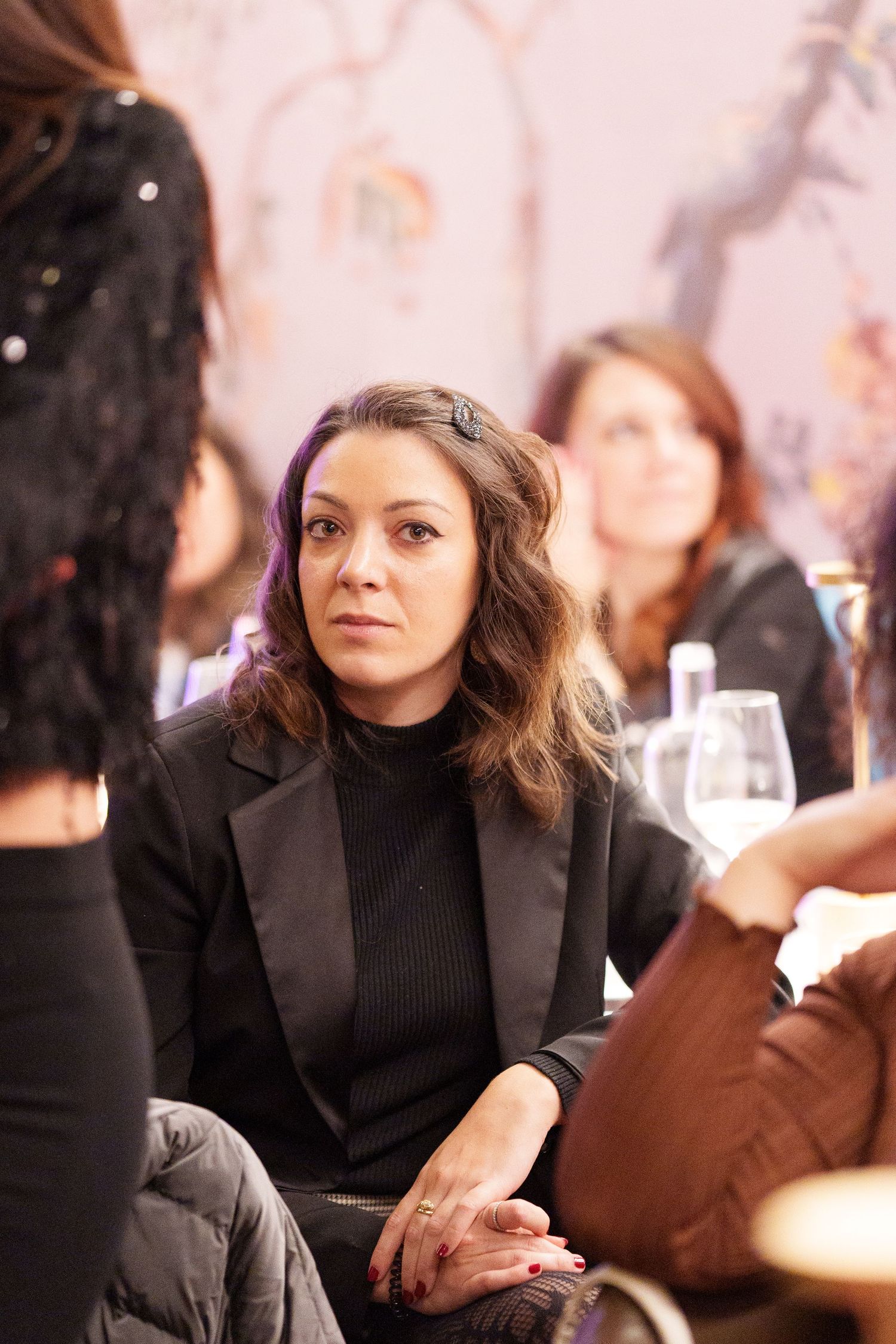 Woman in black blazer sits attentively at an indoor event, with guests and wine glasses visible in the background.