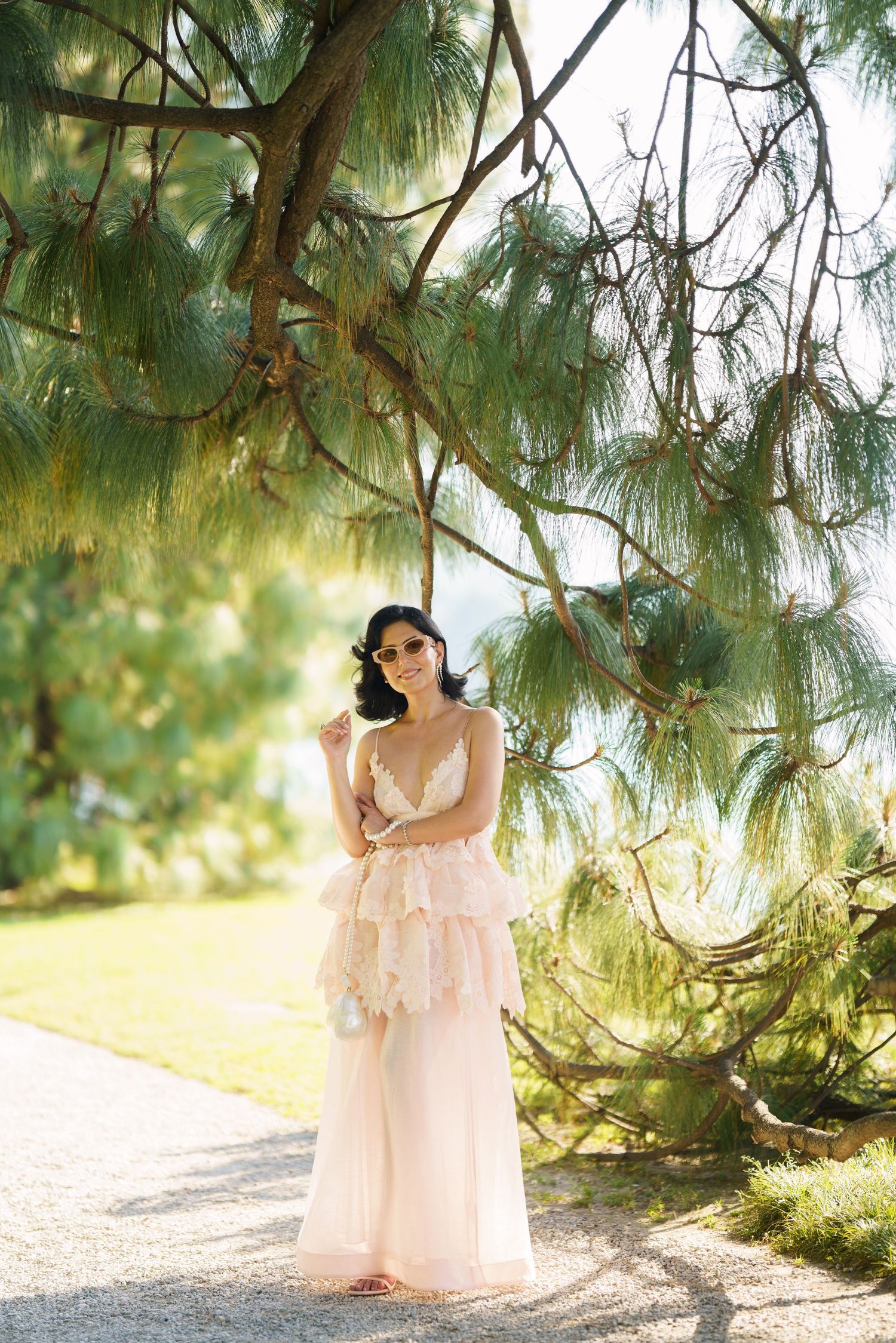 A woman in a blush pink tiered gown poses gracefully beneath pine tree branches in a sunlit garden.