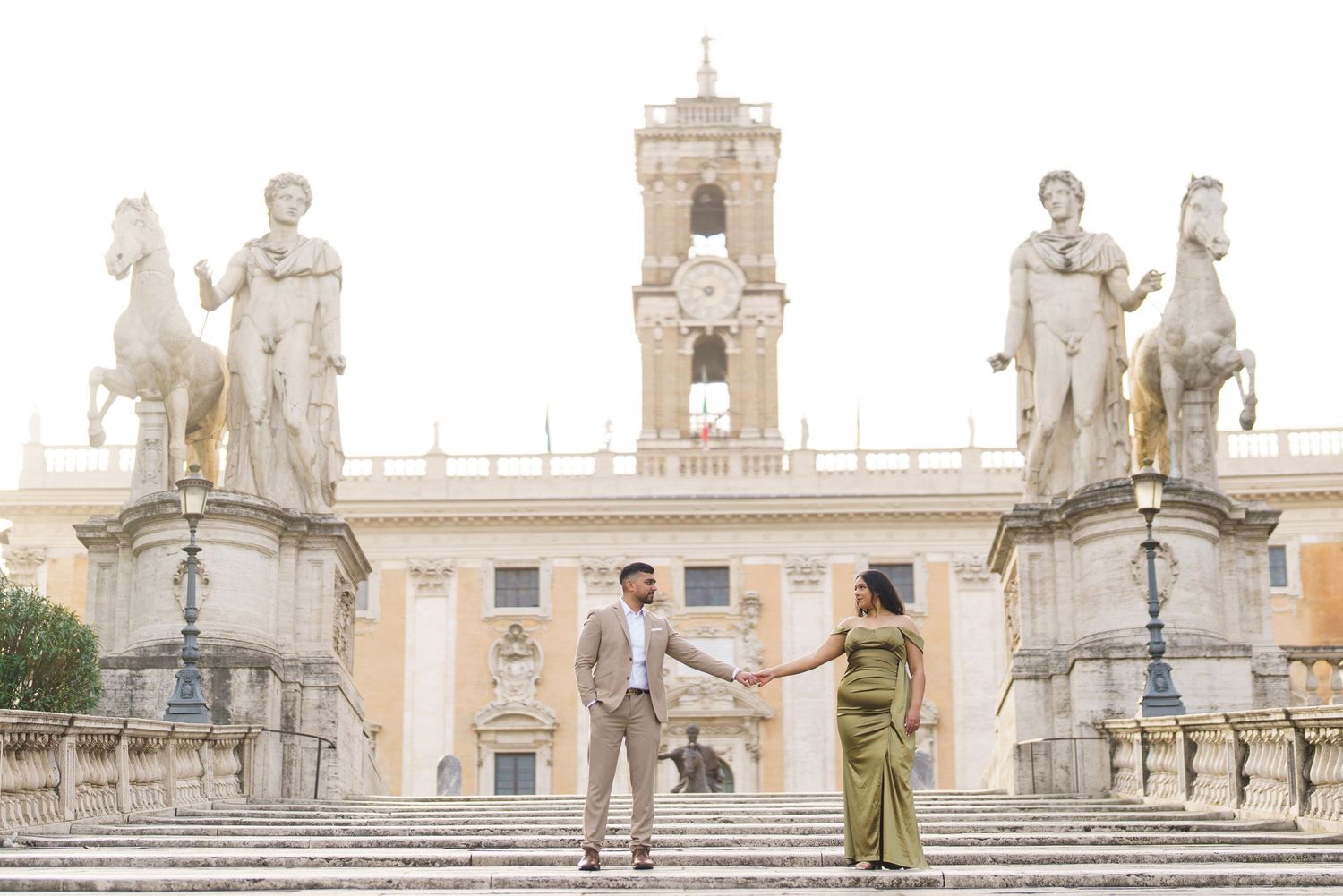 Couple holds hands on Capitoline Hill steps in Rome, with classical statues and bell tower backdrop.