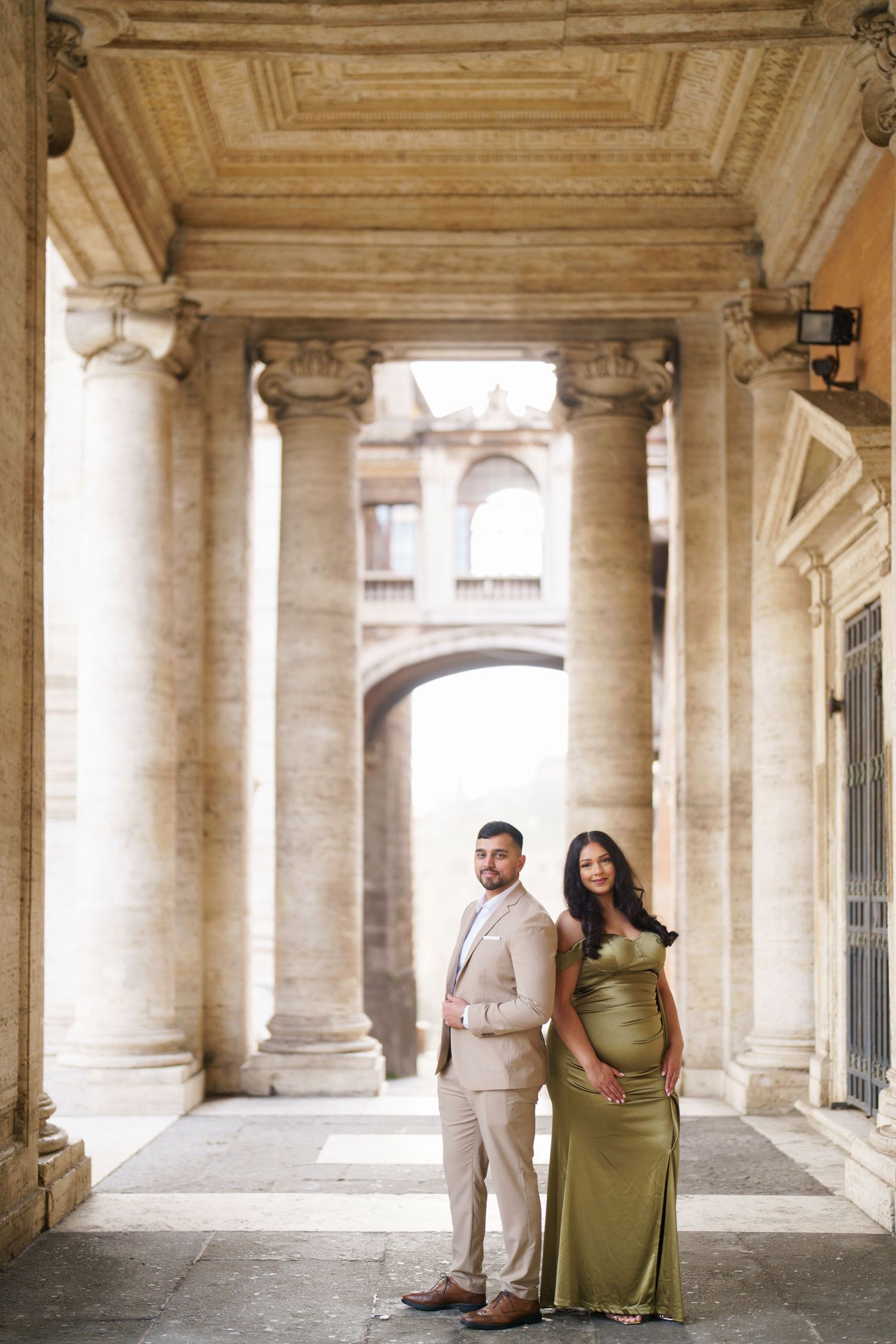 Couple in elegant attire posing under Roman columns, woman in green gown, man in beige suit, classical architecture.