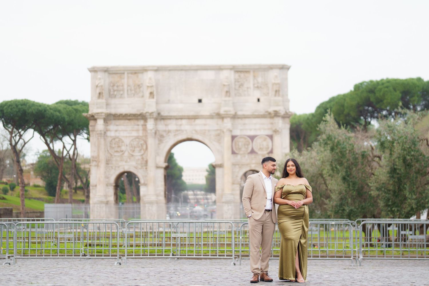 Couple posing in front of the Arch of Constantine in Rome, Italy, with lush green trees in the background.