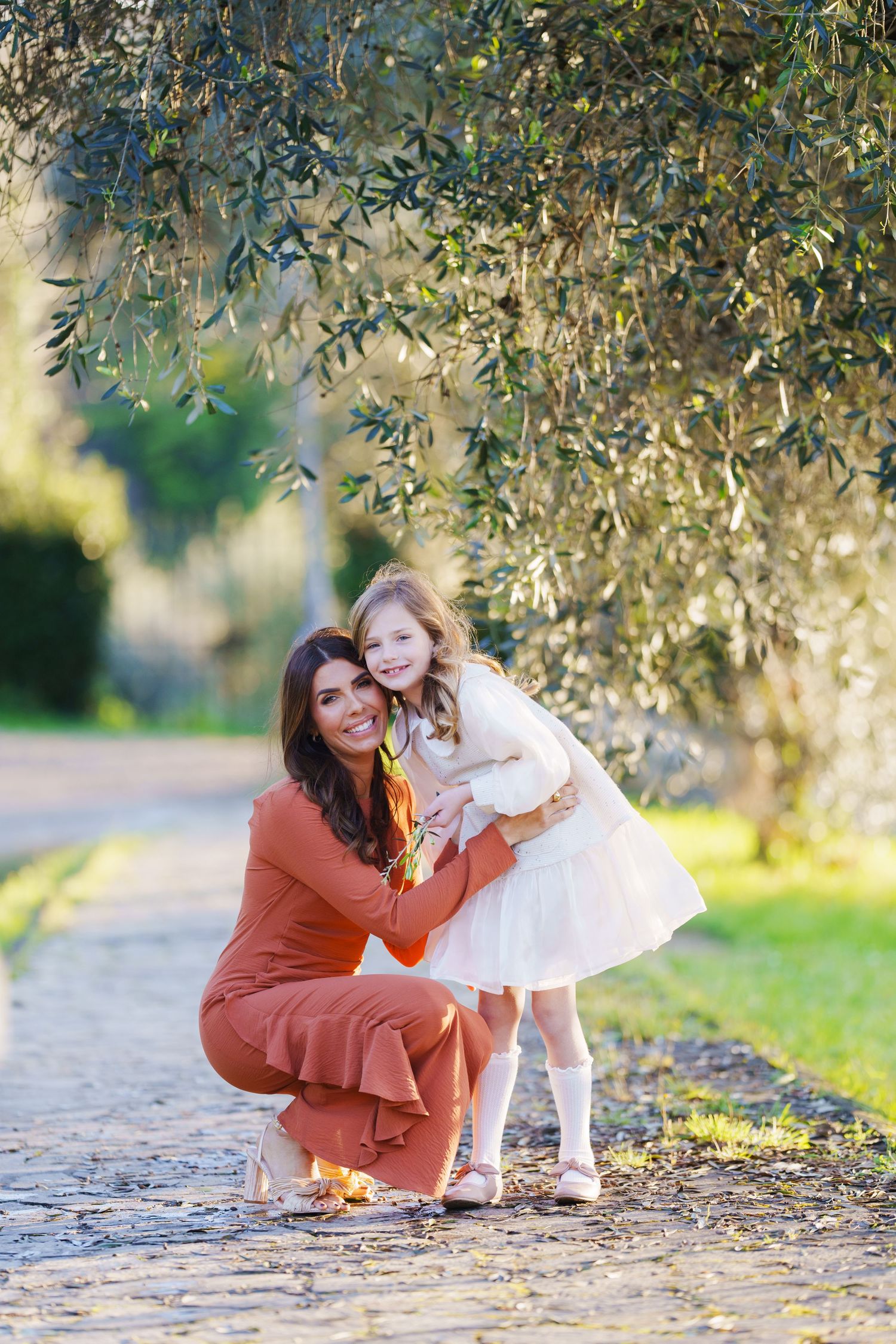 A smiling mother in rust orange outfit crouches to hug her young daughter in white dress outdoors.