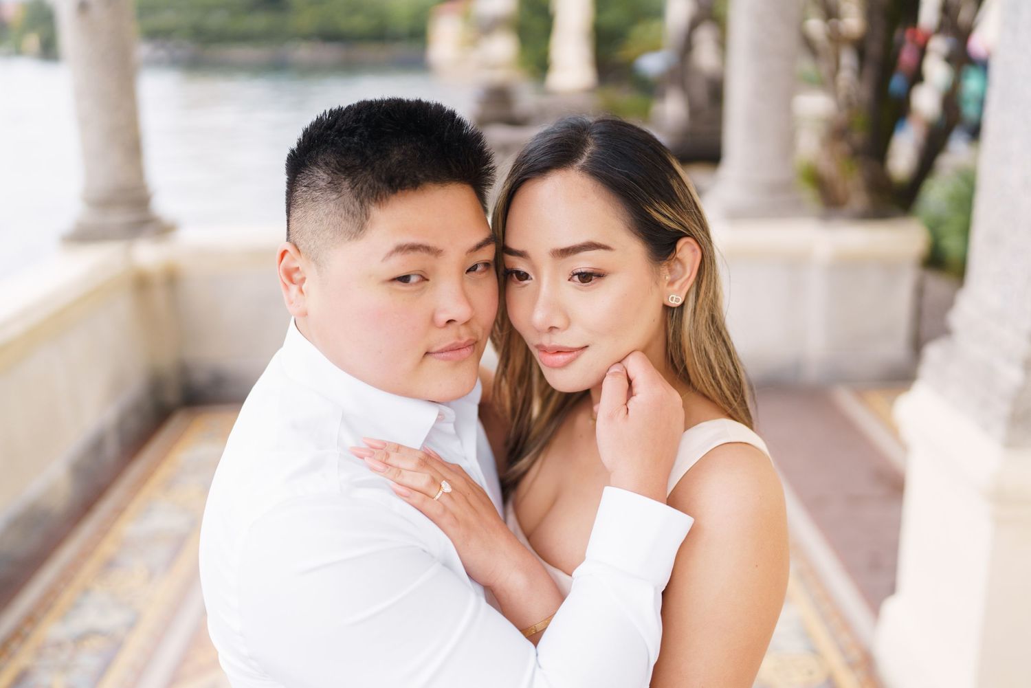 Couple in white attire sharing a romantic embrace outdoors near a waterfront venue.