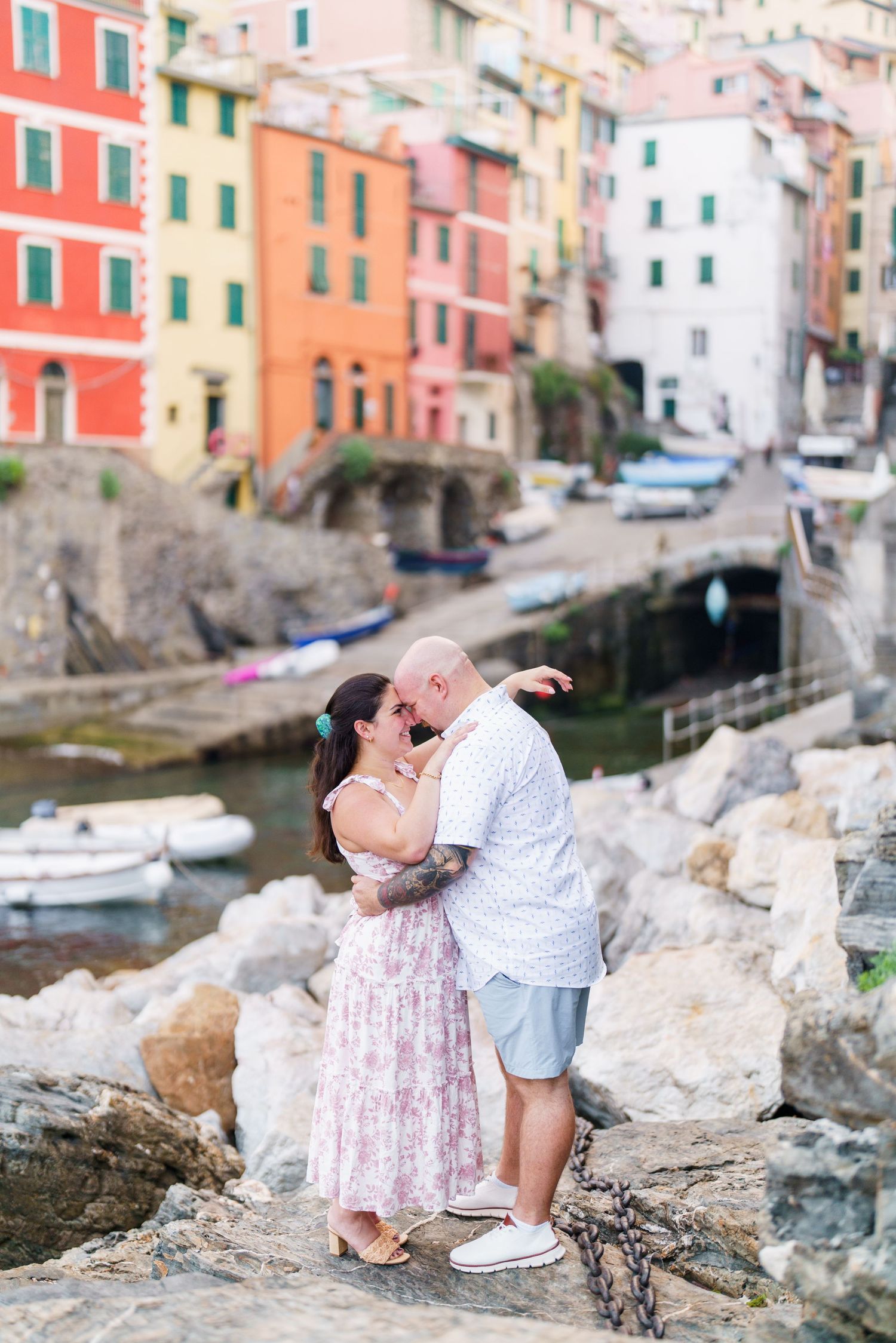 Couple kissing on rocks in Riomaggiore, Cinque Terre, with colorful Italian coastal buildings and harbor in background.