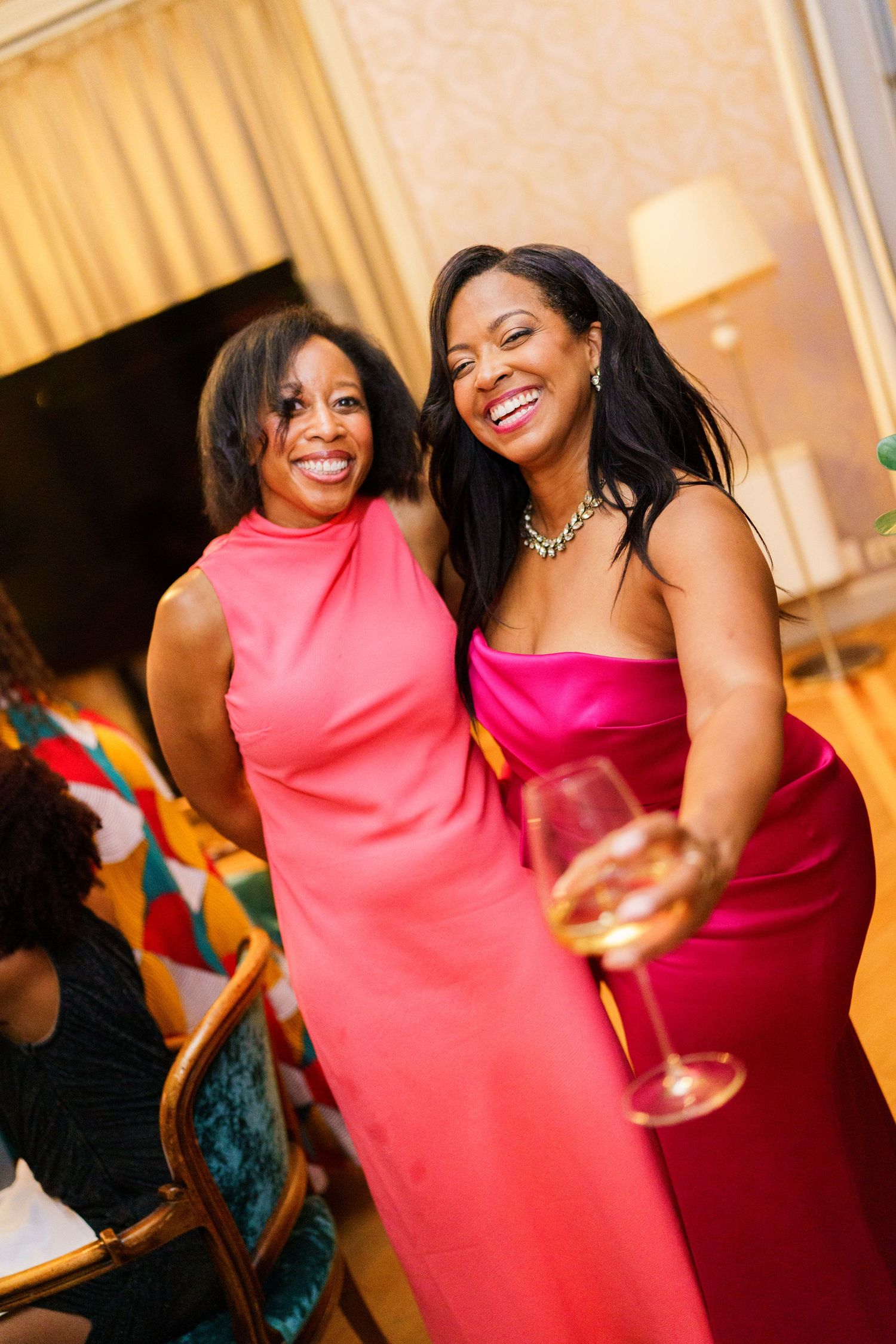 Two women in pink gowns smile and laugh at an elegant event, one holding a wine glass.