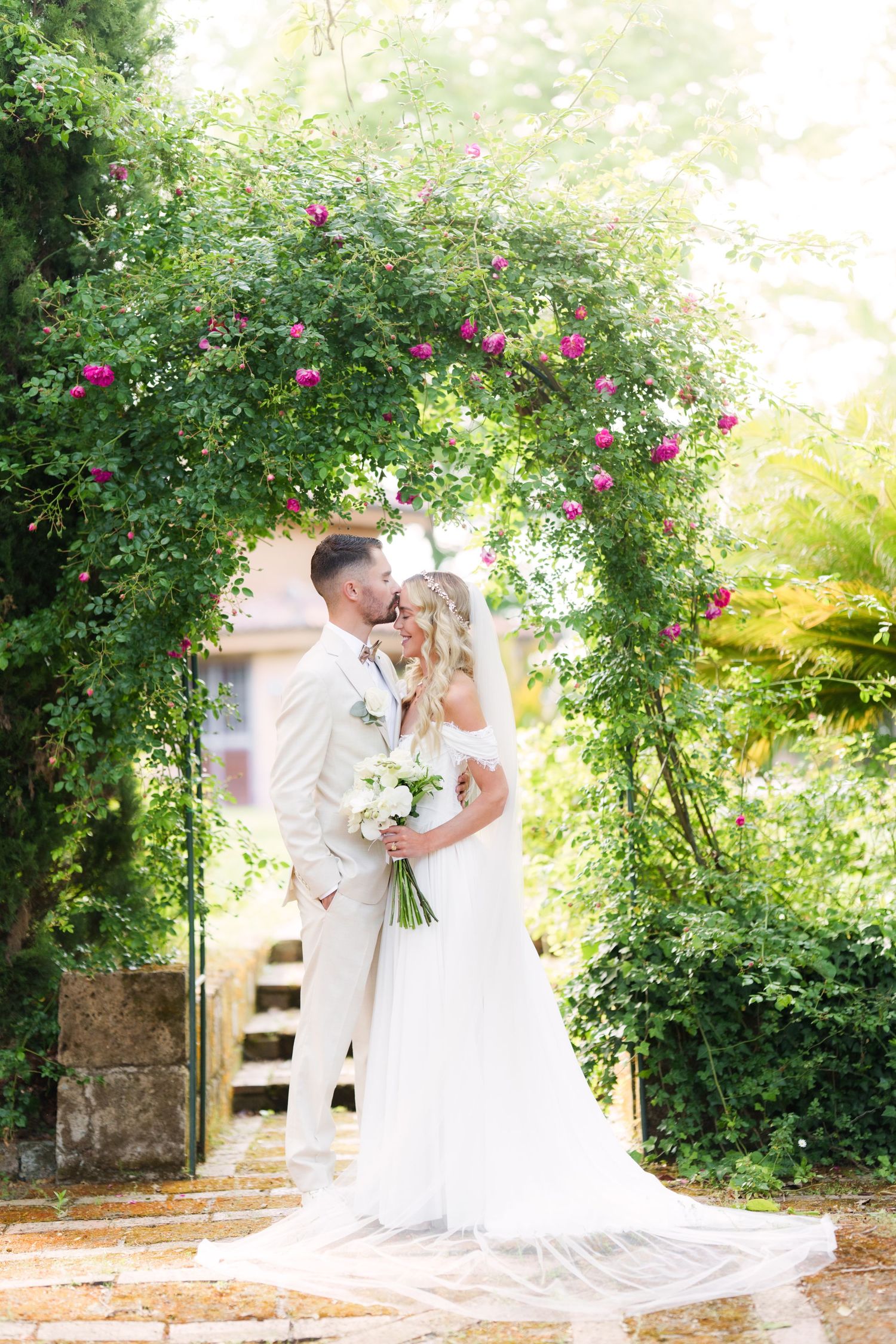 Bride and groom kissing under a floral arch with pink roses and lush greenery on their wedding day.