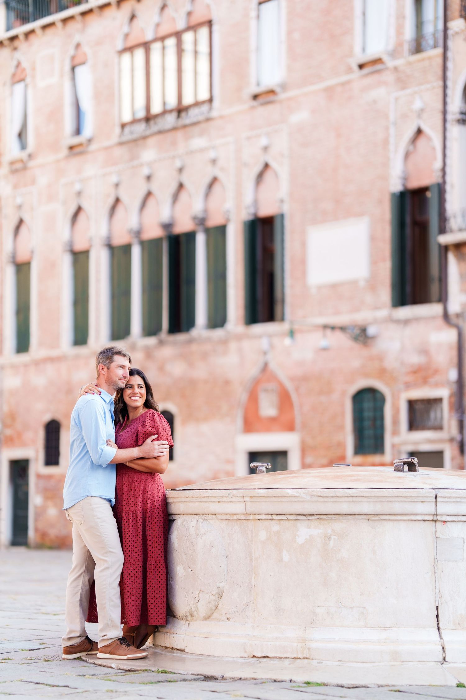 Couple embracing by a stone well in a Venice piazza, with historic red brick architecture in the background.