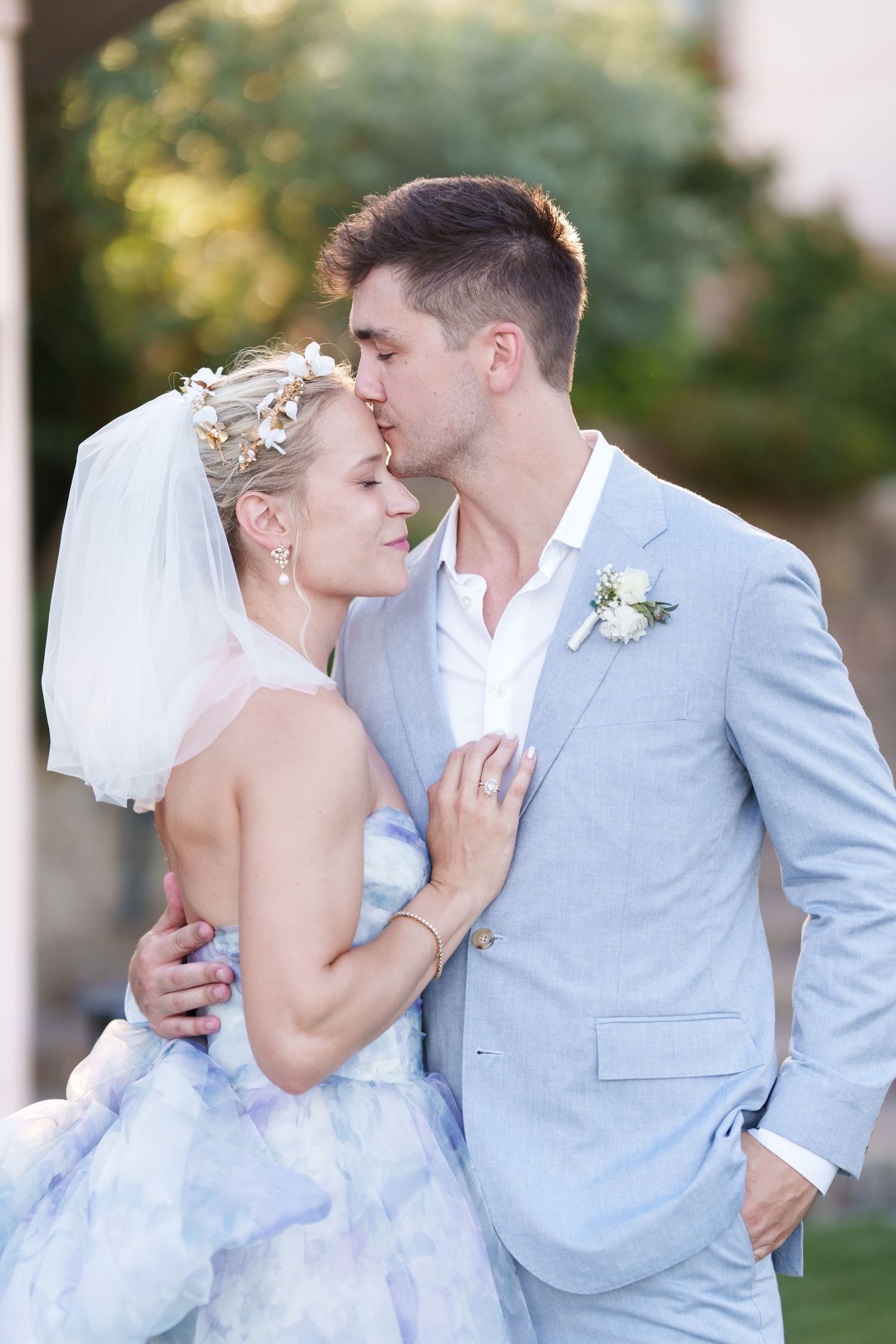 A bride in a floral crown and blue watercolor gown shares a tender moment with her groom in a light blue suit outdoors.