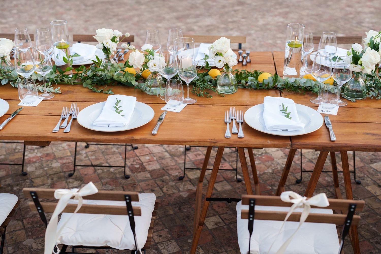 Elegant outdoor wedding reception table with white plates, floral garland centerpiece, and rustic wooden farm tables.